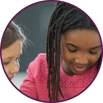 A multi-ethnic group of elementary age children are at their desk in class  Two girls are reading a book together 